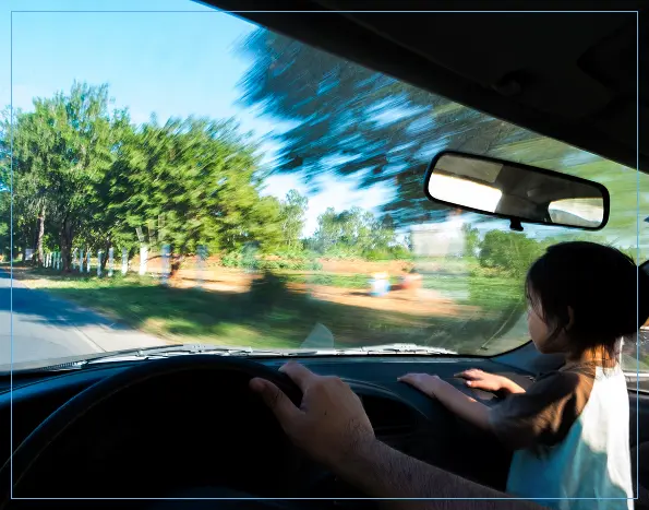 A young child stands unrestrained on the front passenger seat of a moving car, holding onto the dashboard. The driver’s hand is visible on the steering wheel, and the background outside the windshield appears blurred due to motion. The scene suggests a lack of proper child safety measures.